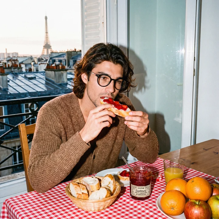 A man at a Paris breakfast table eating jam-topped bread, a green heads-up display in the lens showing steps, battery, and today's task list