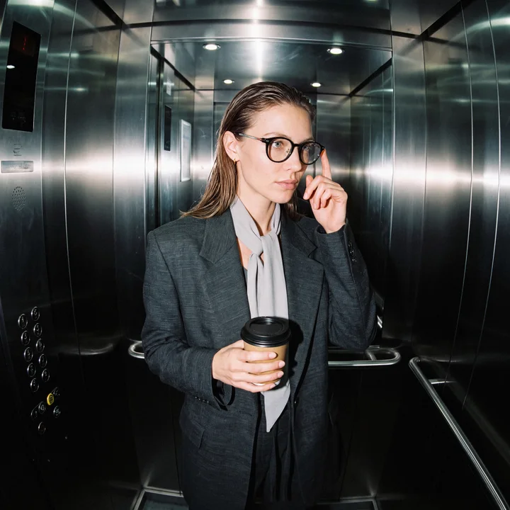 A woman in a steel elevator holding a coffee cup, touching the temple of her glasses as she reads a reply before it is sent