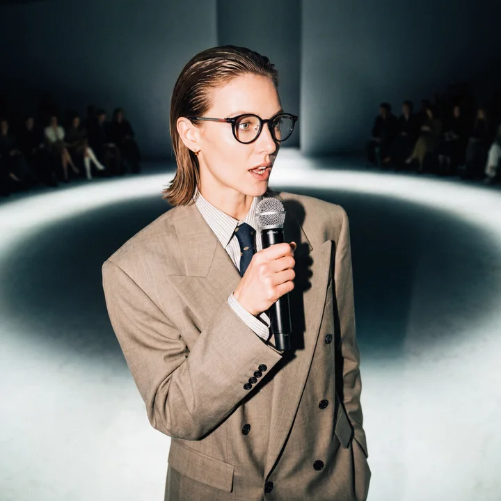 A woman speaking into a handheld microphone on a runway-lit stage, rows of a seated audience visible in the dark on either side